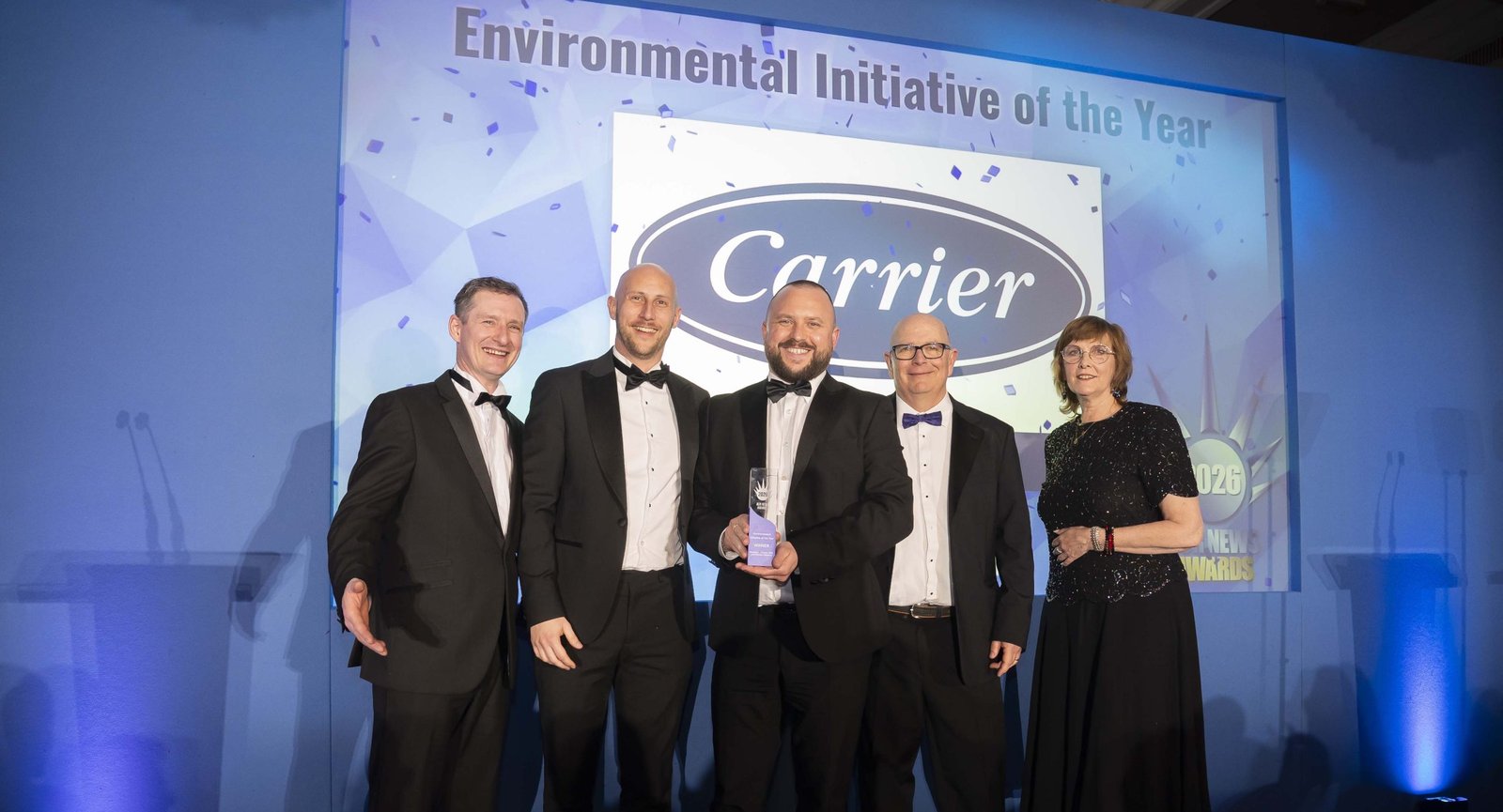 Five people in evening wear on stage beneath a screen reading Environmental Initiative of the Year, with the logo of our supplier partners Carrier, holding the winner's trophy