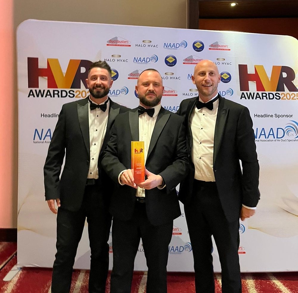 Three men in evening wear in front of an HVR Awards 2025 backdrop, one holding the winner's trophy